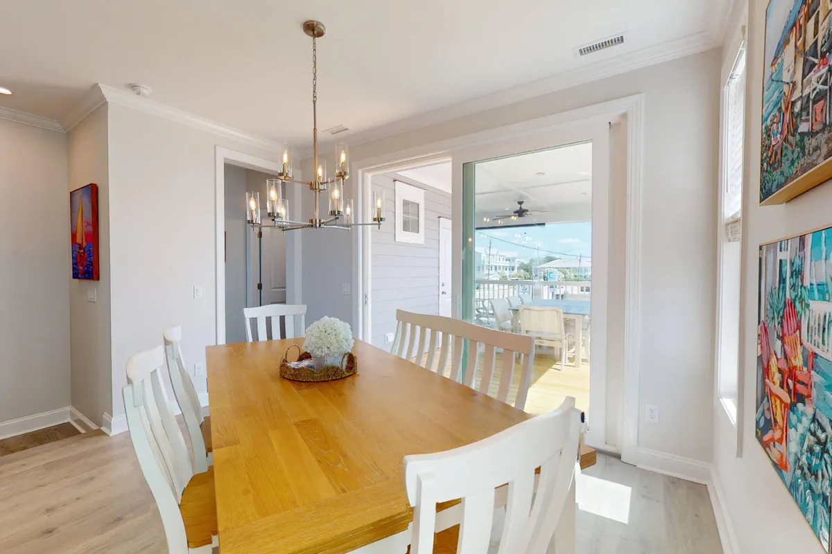 An inviting dining area with a wooden table, elegant chandelier, large glass doors opening to the patio, colorful artwork, and a decorative centerpiece. The space is filled with natural light, offering a direct visual connection to the outdoor deck.