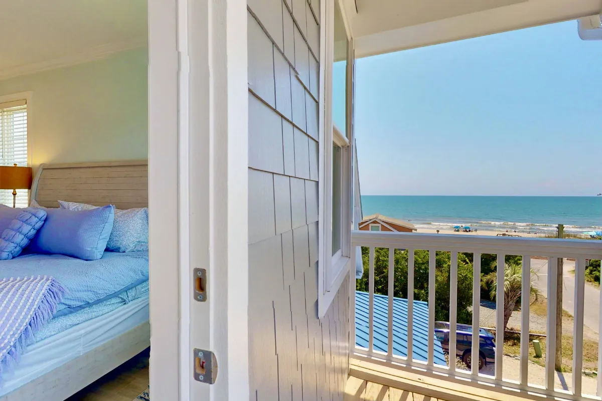 A tranquil bedroom featuring a bed with blue pillows, an open door leading onto a balcony with a clear ocean view, and ample natural light. The relaxing setup hints at opportunities for enjoying the ocean breeze and morning coffee right from bed.