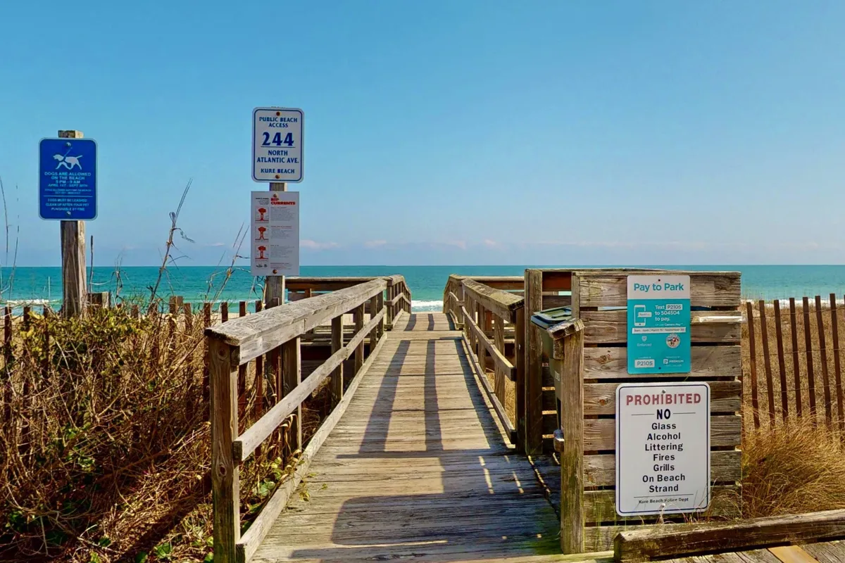 This image captures a wooden boardwalk leading directly to the ocean, with inviting signage, expansive views of the beach, and the water stretching out towards the horizon. The scene is bathed in warm, natural light, emphasizing easy access to the shore and the tranquil, welcoming atmosphere of the property’s surroundings.
