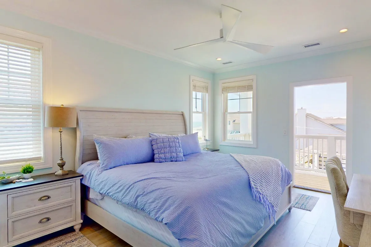 A spacious bedroom with a large bed dressed in blue bedding, a nightstand with a soft lamp, windows filling the room with natural light, and a door that opens directly onto a deck. The inviting ambiance suggests a peaceful retreat ideal for unwinding and enjoying the morning sun.