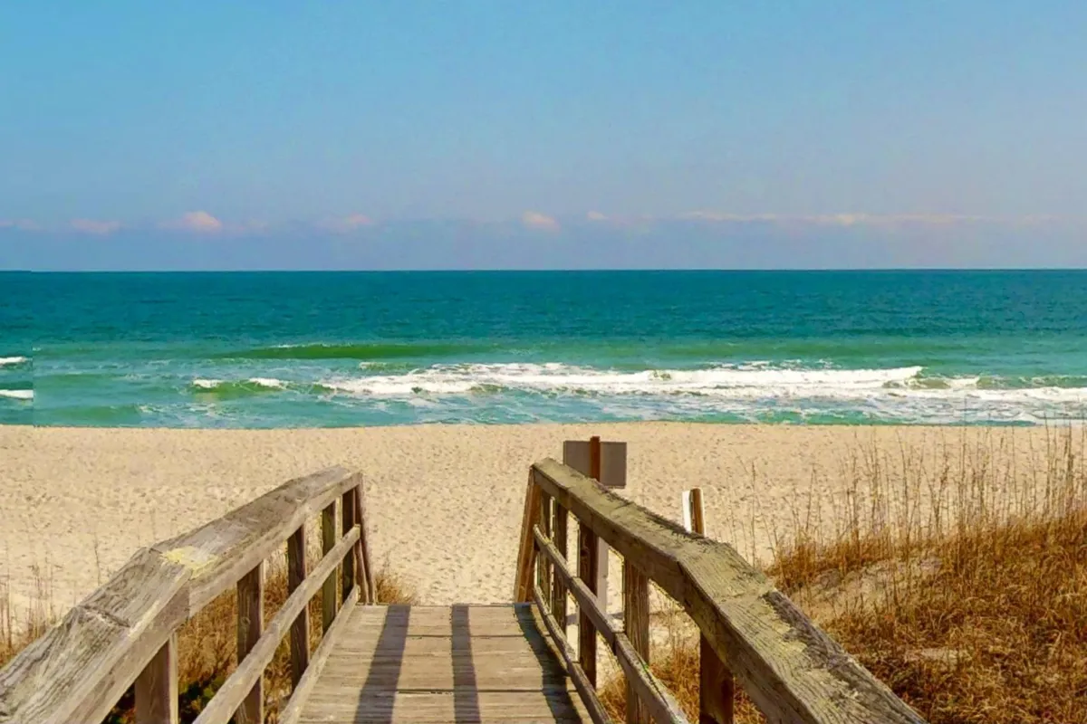 A classic view of a wooden walkway leading directly to the beach, with gentle waves crashing and the open ocean stretching to the horizon. The sandy path is inviting, and the atmosphere is peaceful yet full of potential for adventure, from sunbathing to surfing.