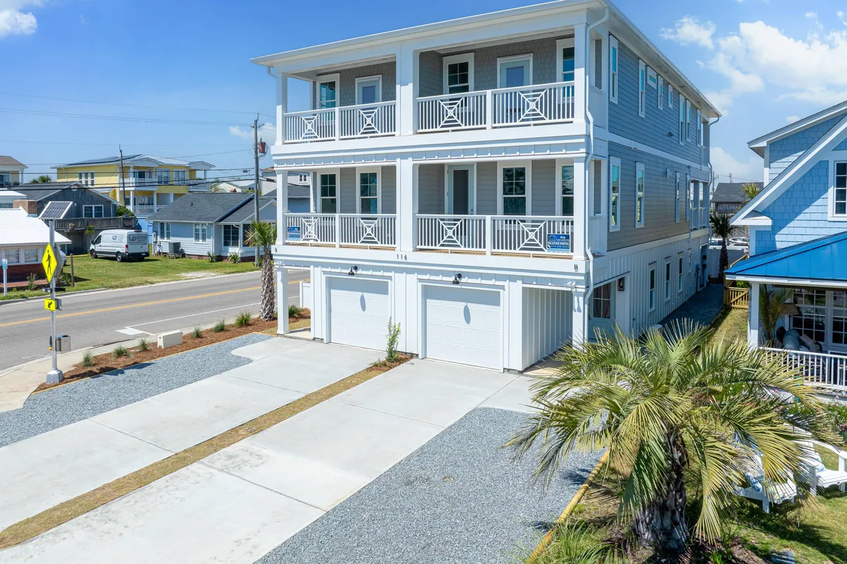 A three-story, modern house stands proudly behind a palm tree, with a wide driveway and a welcoming balcony. The home’s attractive architectural lines and contemporary design suggest a newly built, well-appointed retreat ideal for families or groups. The mood is cheerful and inviting.