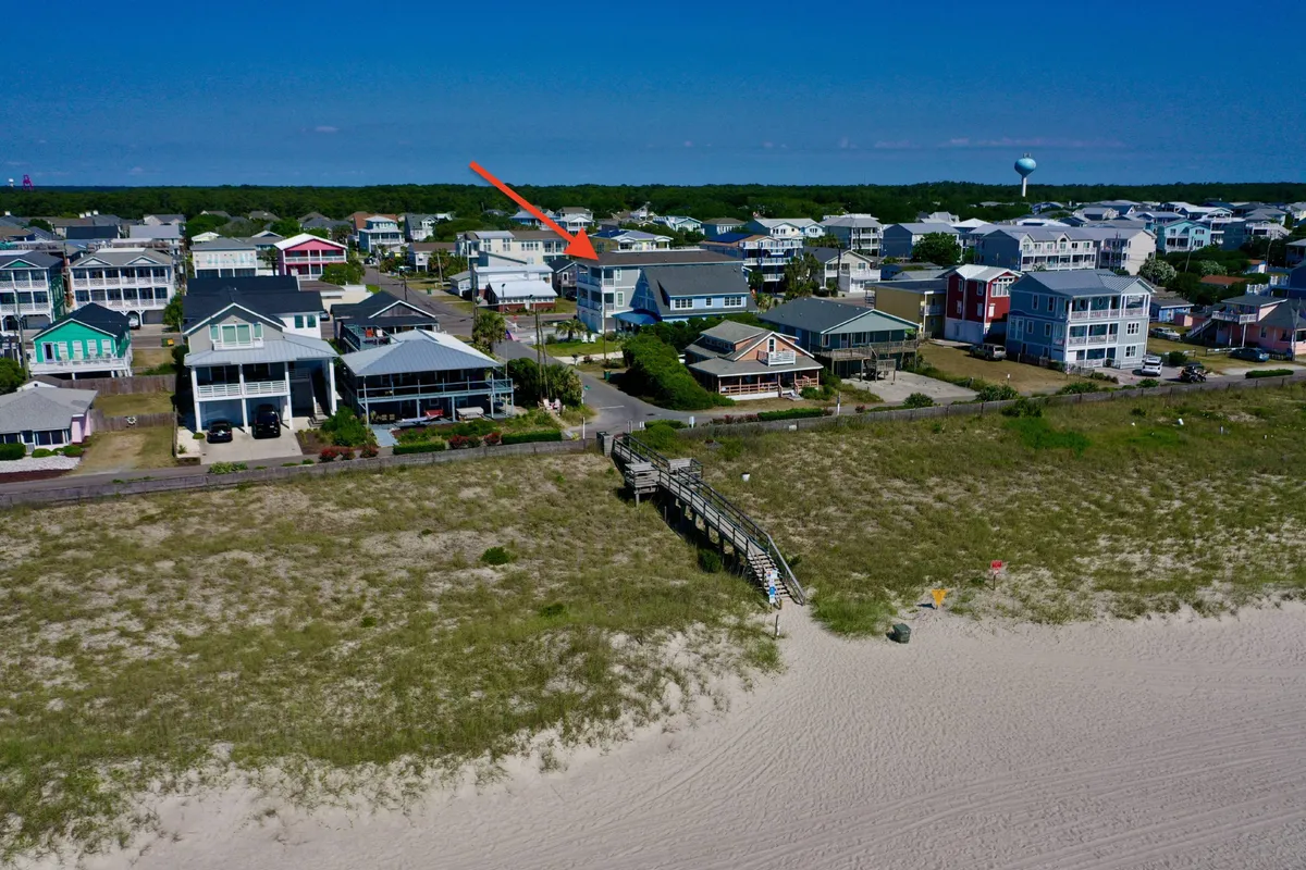 A vibrant exterior view of a beachfront property with a boardwalk leading directly to the ocean. The scene includes surrounding houses and hints at an inviting atmosphere, perfect for gatherings and easy access to the beach. The setting is lively, with the beach just steps away and the suggestion of community and summer events.