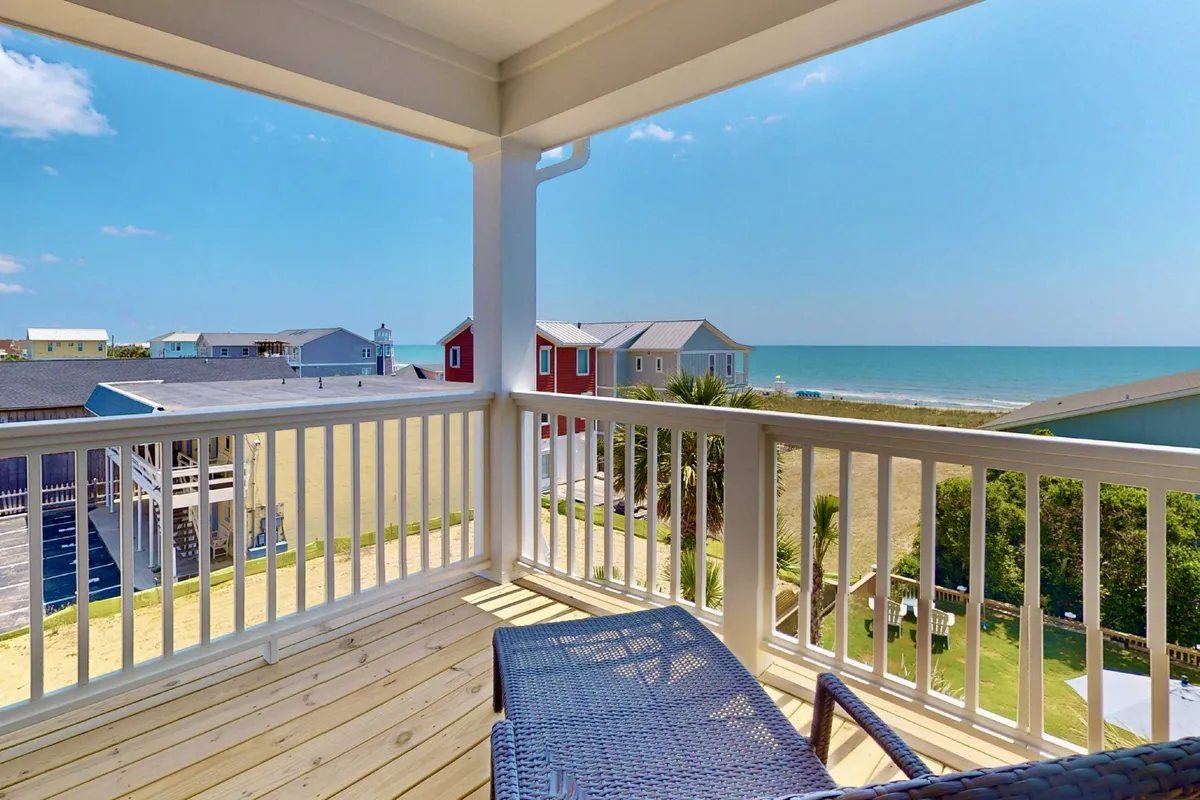 The hero image showcases a serene balcony overlooking sweeping ocean views, framed by a modern railing. Palm trees sway gently in the foreground, with charming beach houses nearby and the inviting shoreline just steps away. The scene invites relaxation and enjoyment of the tranquil coastal atmosphere.