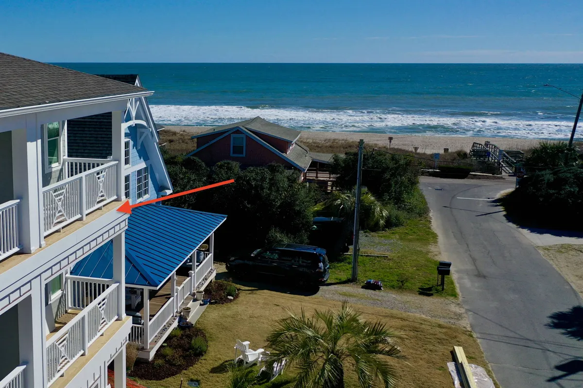 This exterior shot highlights a home with a blue roof and a balcony overlooking the ocean. Palm trees frame the scene, suggesting ample outdoor relaxation and space to enjoy beach views and sunsets together.