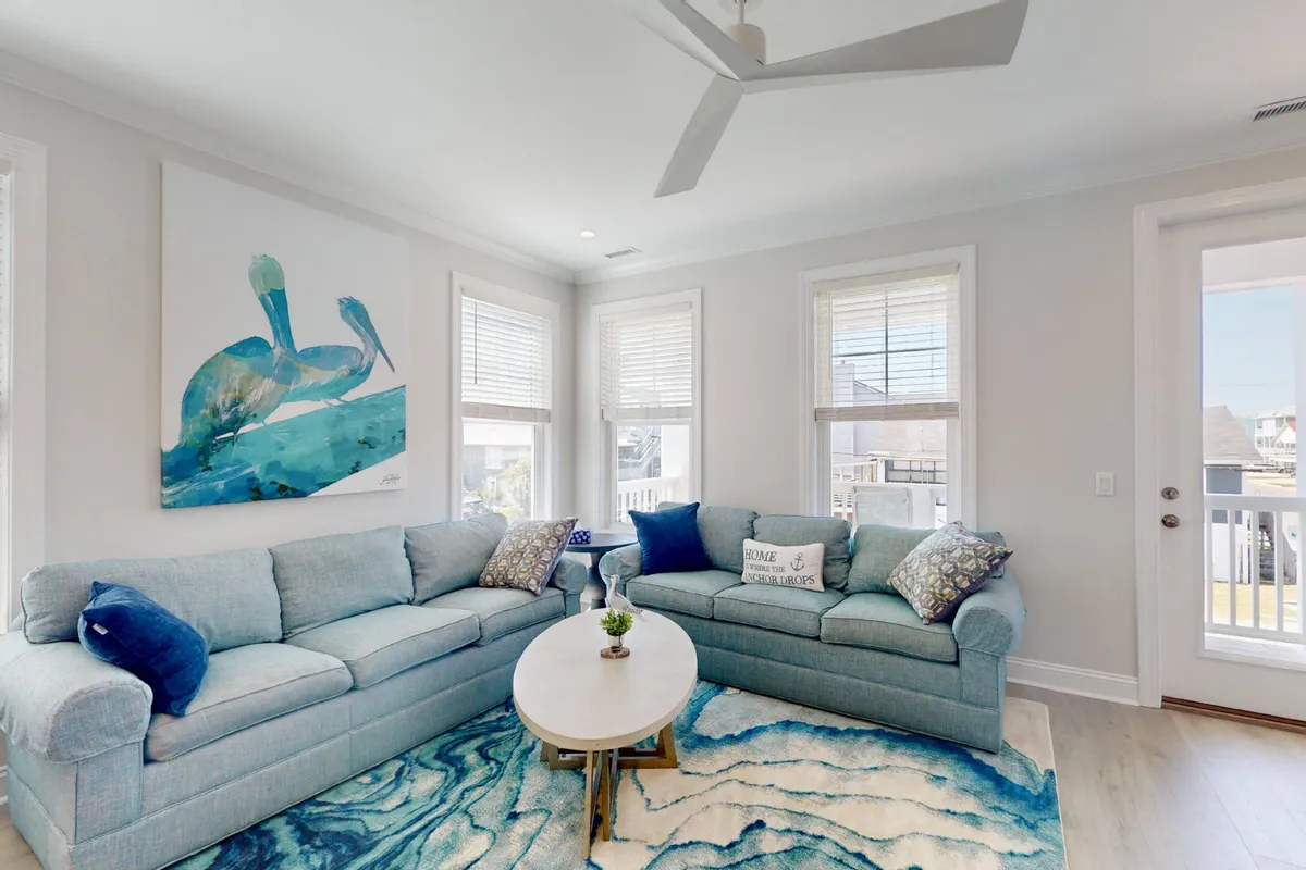 A bright, spacious living room featuring a blue sectional sofa, abstract artwork, a round coffee table, and a colorful area rug. Sunlight floods the space, creating a cheerful and inviting area perfect for relaxation or socializing.