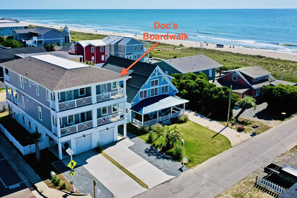 This exterior image features the stretch of beach in front of the property, neighboring houses, the recognizable Doc's Boardwalk sign, and the sparkling ocean beyond. The scene includes people walking on the sand, groups gathering, and hints of local businesses nearby, all framed under the warm light of either sunrise or sunset.