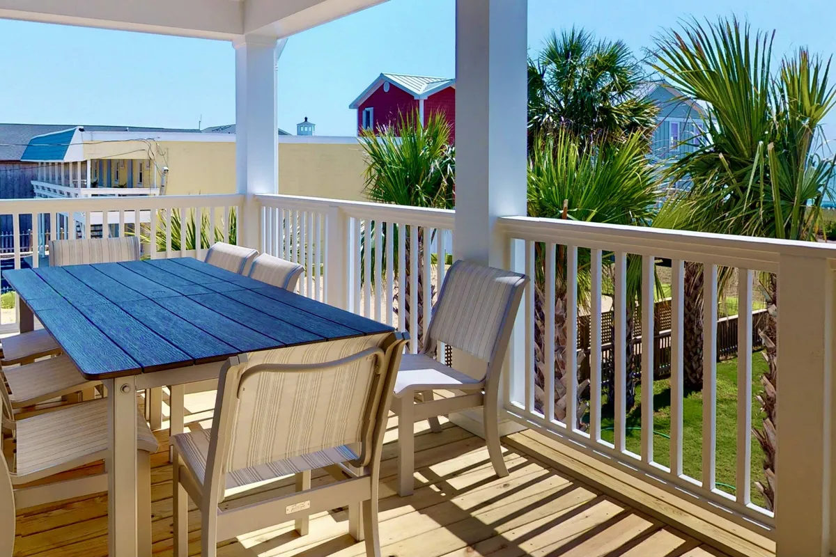 A cheerful outdoor dining area featuring a wooden table with white chairs surrounded by lush green palm trees. The setup overlooks an open, sunlit space, ideal for group meals and social gatherings. The atmosphere is bright and inviting, perfect for enjoying meals with a view of the outdoors.