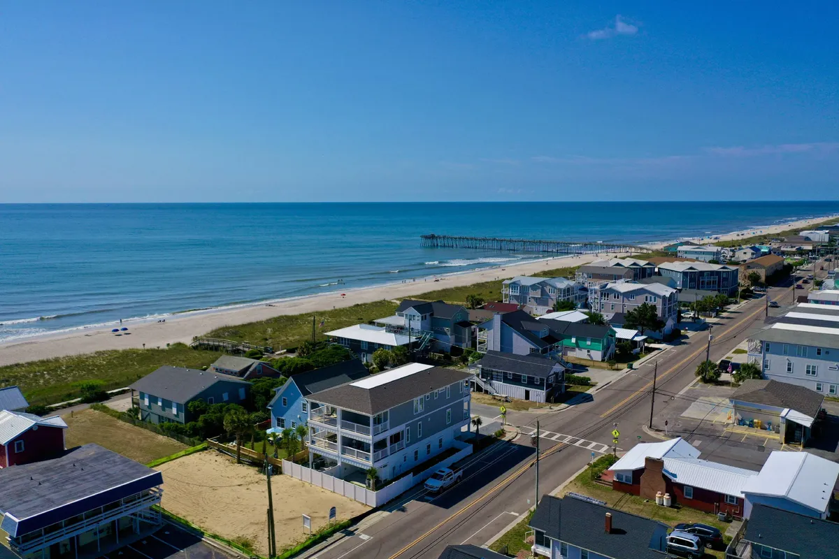 A scenic view encompassing the beach, Kure Beach Pier, ocean waves, nearby houses, and the adjacent road. The composition captures families strolling, beachgoers relaxing, and the iconic pier stretching out into the water, illustrating easy access to classic seaside attractions.