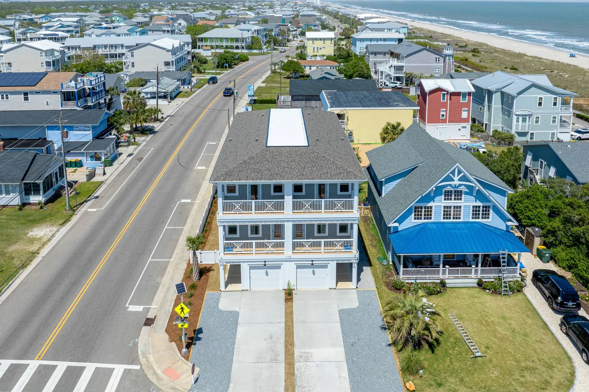 A relaxed exterior shot of a two-story house with a spacious front porch, neighboring homes, and a road leading directly to the beach. The inviting porch hints at sunset views and easy morning access to the beach and local dining.